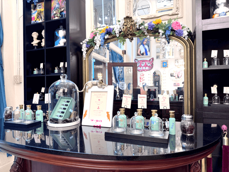 A mirrored vanity displays perfume bottles, testers, and informational signs in a boutique setting, with floral decor on the mirror and shelves of assorted bottles crafted by Asheville artisans in the background.