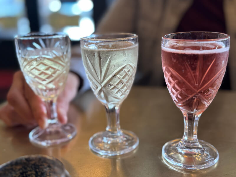 Three crystal glasses containing different colored beverages—white, clear, and pink—are placed on a table, with a hand holding the glass on the left.