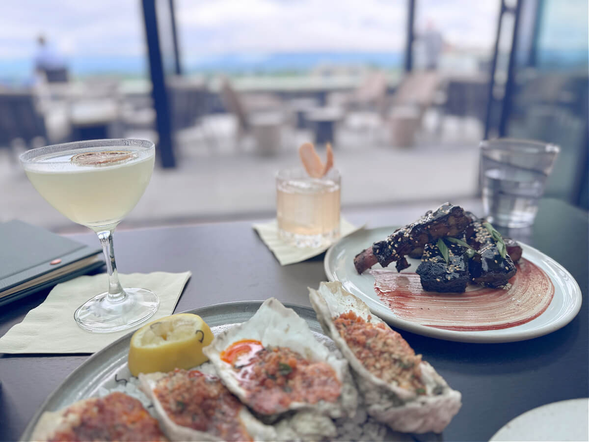 A table at an outdoor Asheville restaurant features a cocktail, a short drink, a plate of seasoned oysters with lemon, a serving of ribs with garnish, and a glass of water.