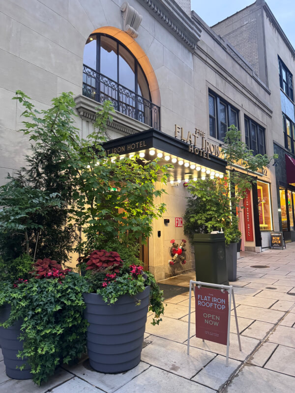 Entrance of The Flat Iron Hotel with marquee lights, potted plants, and a sign advertising the rooftop. The sidewalk and neighboring storefronts are visible.