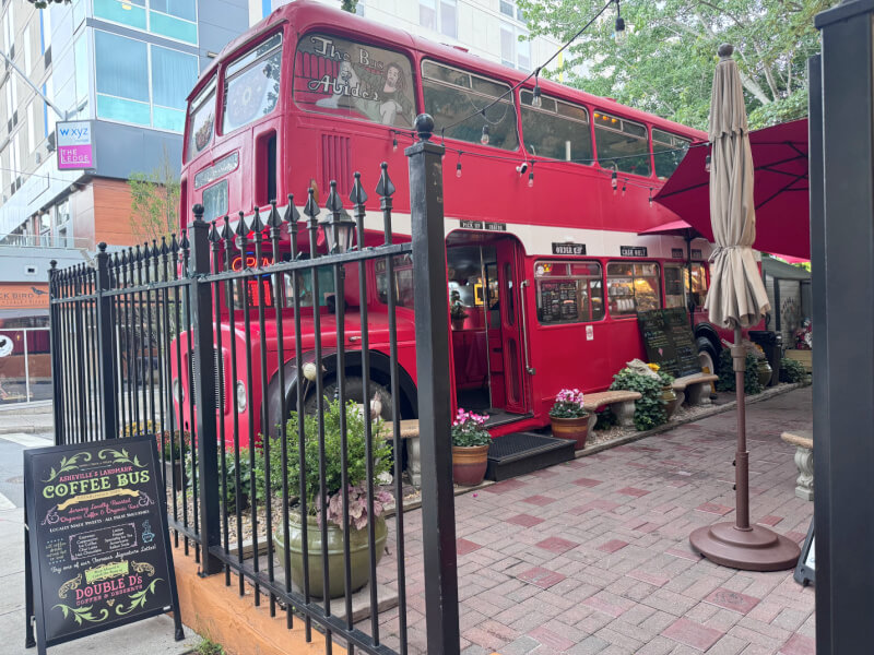 A red double-decker bus converted into a café, one of the most unique Asheville restaurants, sits behind a black fence on a brick patio with potted plants and an outdoor sign displaying the menu.