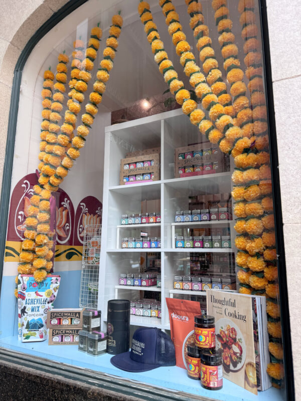 A shop window display featuring colorful spice jars, cookbooks, canned goods, a navy hat, and yellow flower garlands hanging along the glass.