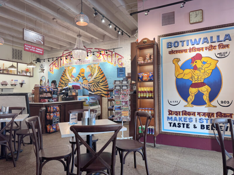 Interior of a casual Asheville restaurant with Indian-themed decor, featuring colorful murals, a counter area, shelves with snacks, and several empty tables and chairs.