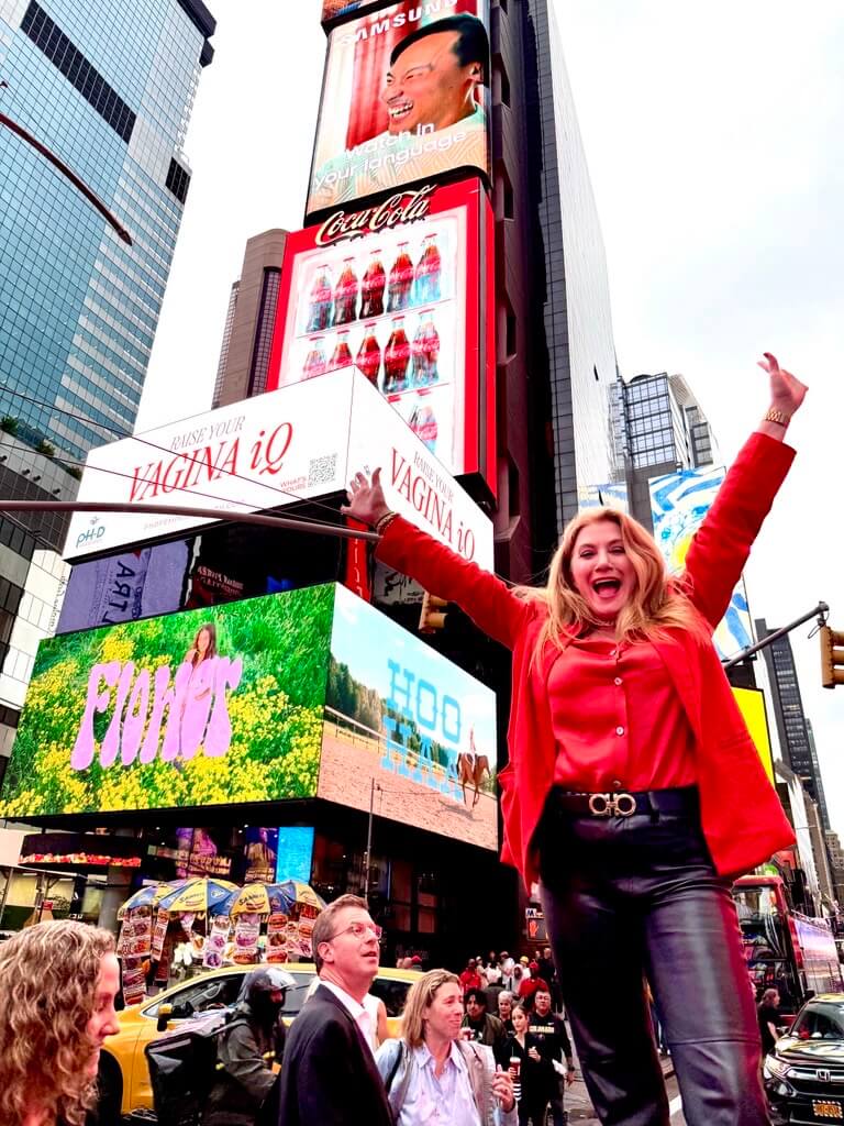 A woman in a red shirt and black pants stands with arms raised in Times Square, surrounded by crowds and colorful electronic billboards, embodying the vibrant energy.