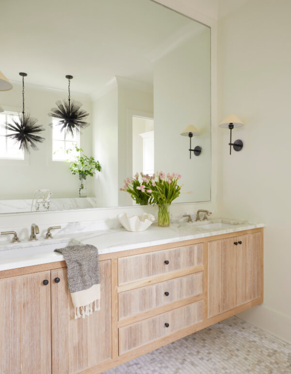 A modern bathroom by Logos Interiors features a light wood vanity, marble countertop, double sinks, a large mirror, wall sconces, a gray towel, a vase of flowers, and natural light streaming in from the window.