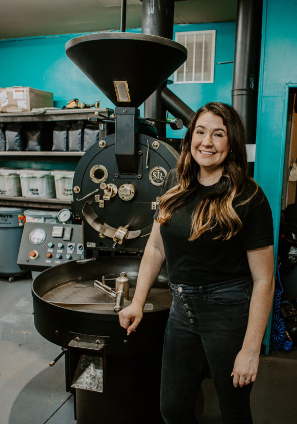 A woman stands smiling next to a large coffee roasting machine in an industrial workspace, with bags of Summit Sisters Coffee beans on shelves in the background.