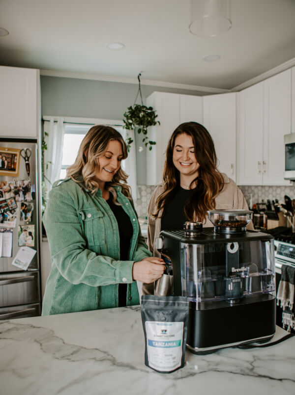 Two women stand in a kitchen using an espresso machine on a marble countertop, with a bag of Summit Sisters Coffee Tanzanian beans in front of them.