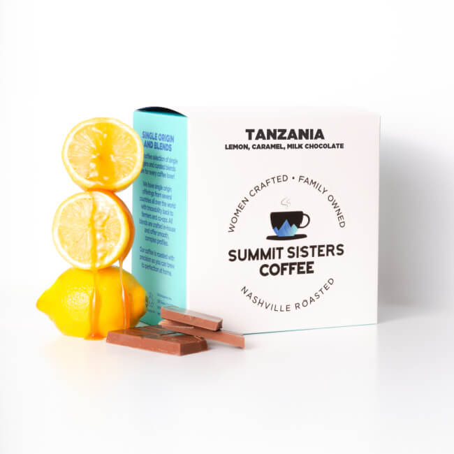 A box of Summit Sisters Coffee sits beside a halved lemon, a whole lemon, and two pieces of milk chocolate on a white background—perfect for those who enjoy unique coffee pairings.