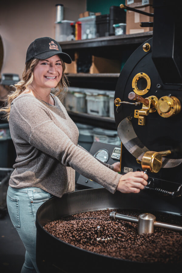 A woman wearing a cap smiles while operating a large coffee roasting machine filled with beans in an industrial setting at Summit Sisters Coffee.