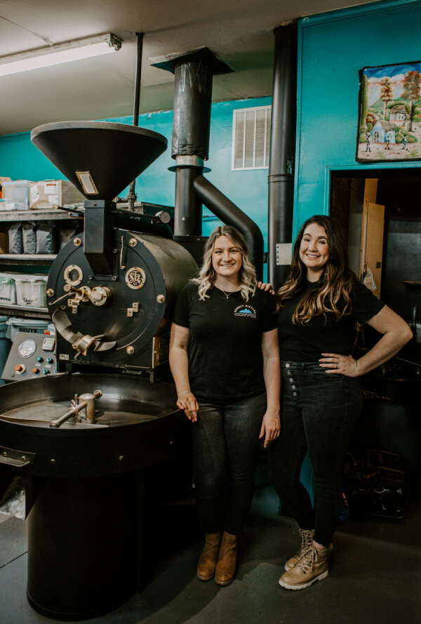 Two women stand next to a large Summit Sisters Coffee roasting machine in an industrial-style room with teal walls and packaged goods on shelves in the background.