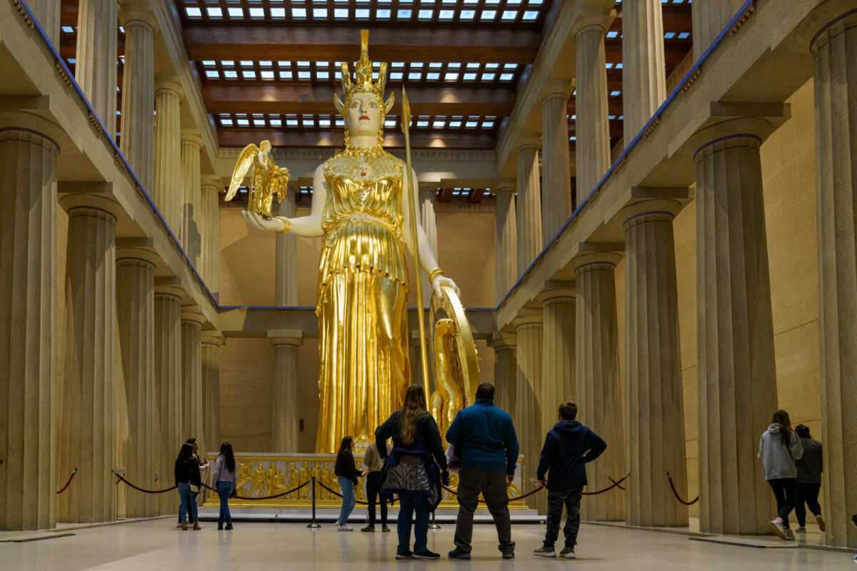 Meet Nashville's Own Golden Goddess: Athena Parthenos - 1 Visitors stand in front of a large gold statue of Athena inside Nashville’s Parthenon, surrounded by tall columns and a skylight ceiling.