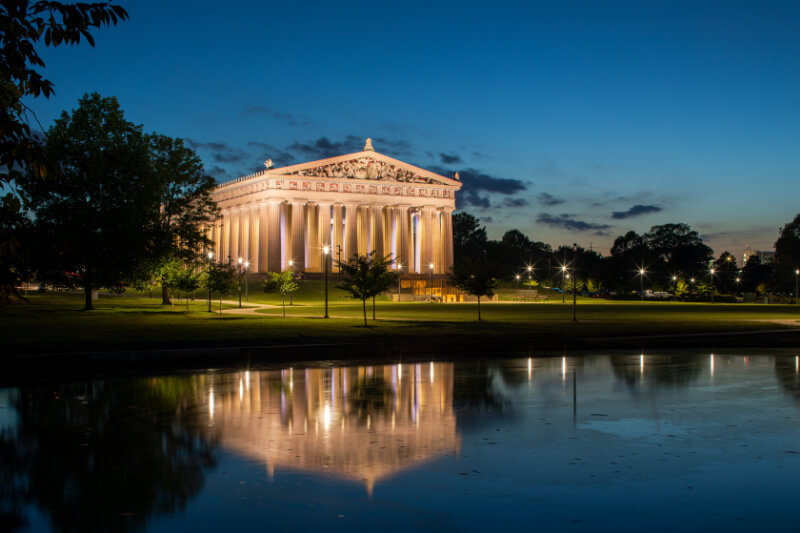Meet Nashville's Own Golden Goddess: Athena Parthenos - 2 The Parthenon replica in Nashville, Tennessee, dedicated to Athena, is illuminated at dusk, its reflection shimmering in a calm pond in the foreground.
