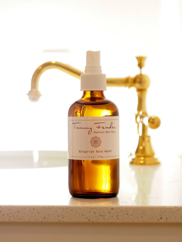 An amber spray bottle of Tammy Fender Holistic Skin Care Bulgarian Rose Water sits on a countertop, with a gold faucet in the background.