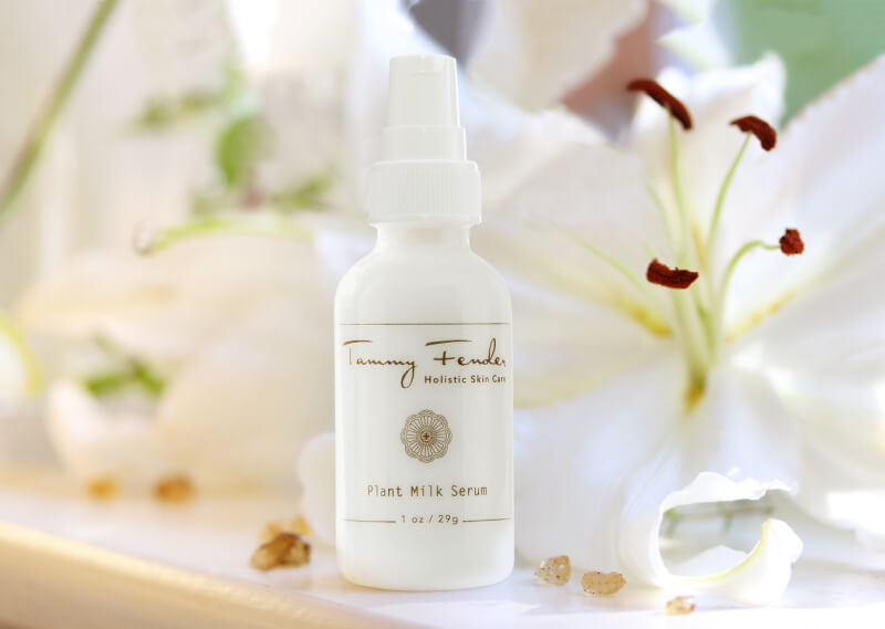 A white bottle labeled "Tammy Fender Plant Milk Serum" stands elegantly before white lilies, bathed in soft natural light.