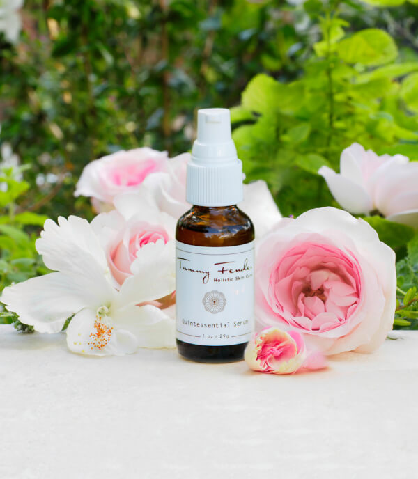 A brown glass dropper bottle labeled "Quintessential Serum" by Tammy Fender stands on a white surface, surrounded by pink and white flowers with green foliage in the background.