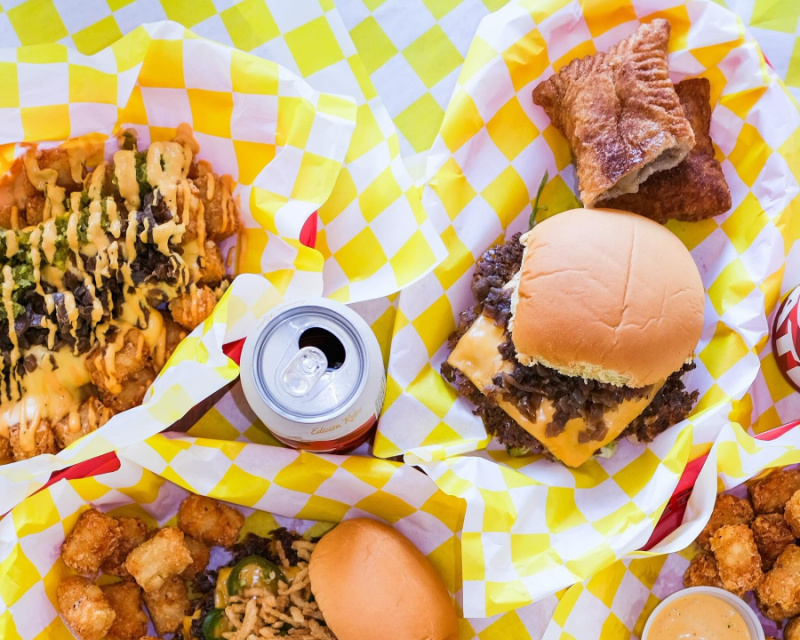 9 New Nashville Restaurants to Try This Month - 1 A meal with a double cheeseburger, crinkle-cut fries with sauce, tater tots, a canned drink, and a fried dessert on yellow and white checkered paper.