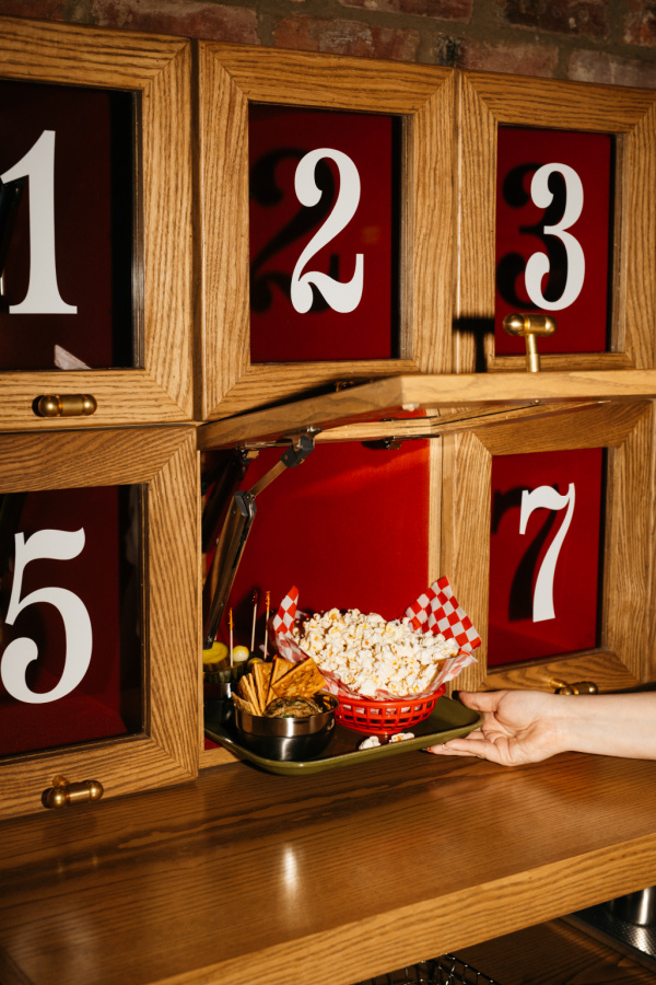 9 New Nashville Restaurants to Try This Month - 2 A hand retrieves a tray with popcorn and snacks from a numbered wooden compartment with red backgrounds, resembling a cubby or locker system.