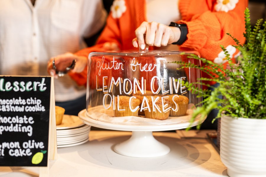 At one of the top new Nashville restaurants, a person in an orange sweater lifts a glass dome labeled "gluten free lemon olive oil cakes" over several small cakes on a white cake stand beside a dessert menu and potted plant.