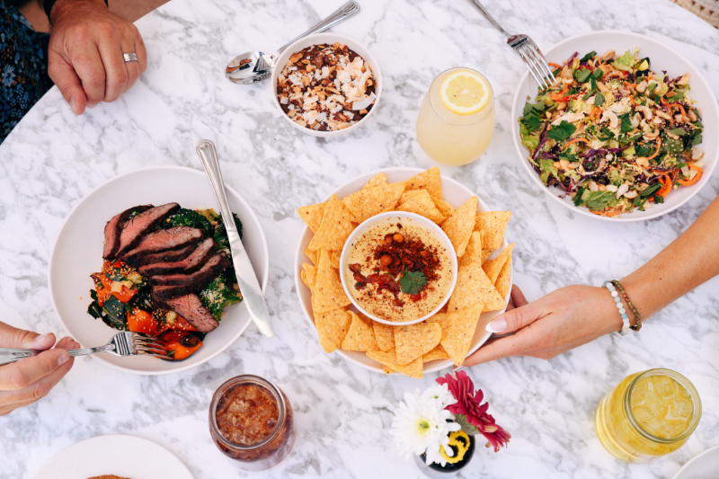 9 New Nashville Restaurants to Try This Month - 3 A marble table set with salad, steak and vegetables, nachos with dip, lemon drinks, a soda, and hands reaching for food at one of the new Nashville restaurants in June 2025.