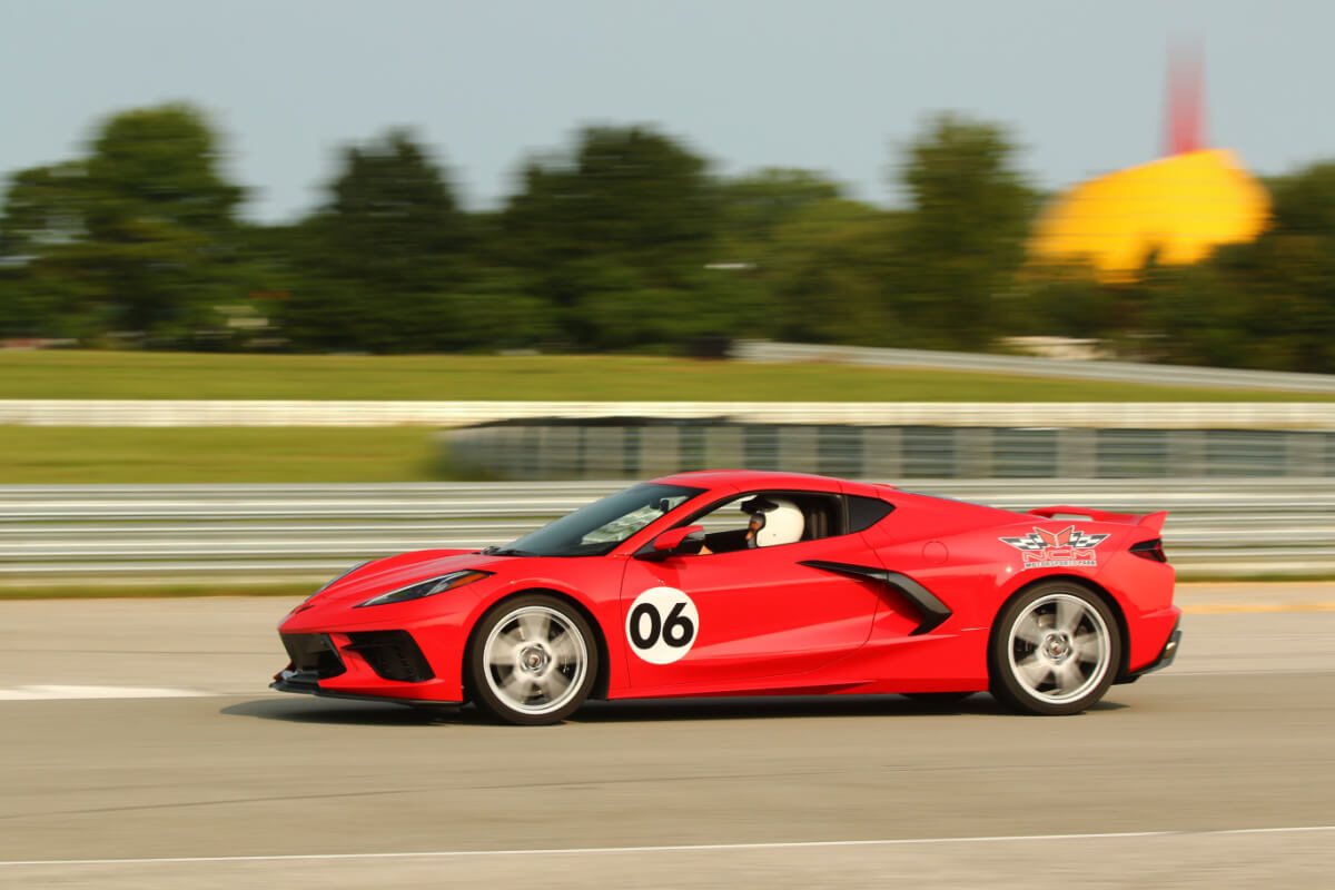 A red sports car with the number 06 on the side speeds along a Nashville race track, with blurred trees and a yellow structure in the background—a perfect scene for summer weekend trips.