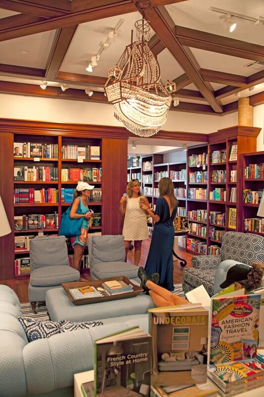 Three women stand and talk in a cozy, well-lit 30A bookstore lounge furnished with blue chairs and sofas, surrounded by bookshelves and a chandelier overhead.