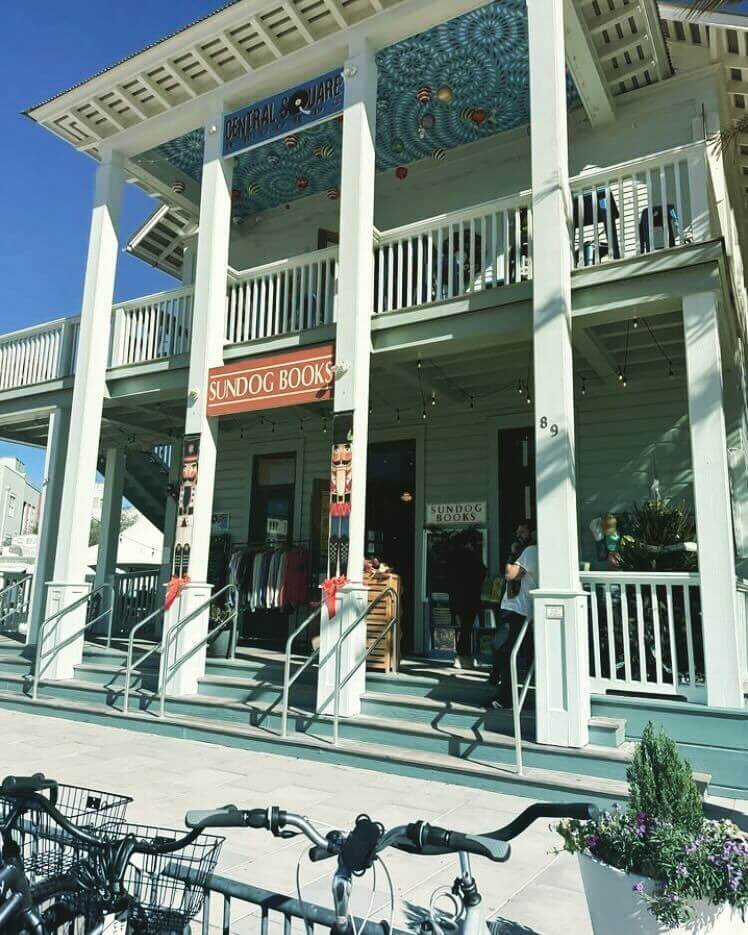 Two-story building along 30a with a sign reading "Sundog Books" on the first floor, a balcony above, and bicycles parked in front on a sunny day.