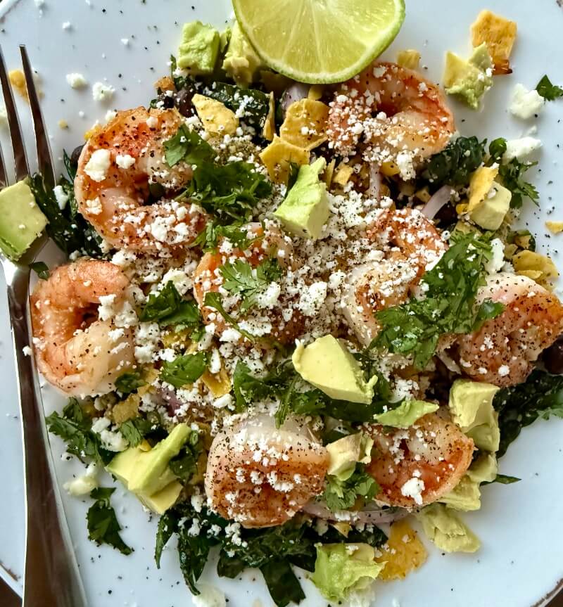 A plate of Shrimp Kale Salad topped with avocado chunks, crumbled cheese, herbs, and a lime wedge, served with a fork and drizzled with tangy Lime Pepita Dressing.
