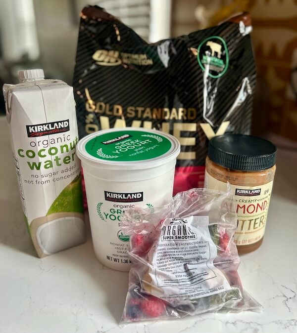A protein powder bag, a carton of coconut water, a tub of Greek yogurt, a jar of almond butter, and a bag of frozen mixed berries—perfect Protein Picks at Costco—sit on the kitchen counter ready for your next healthy creation.