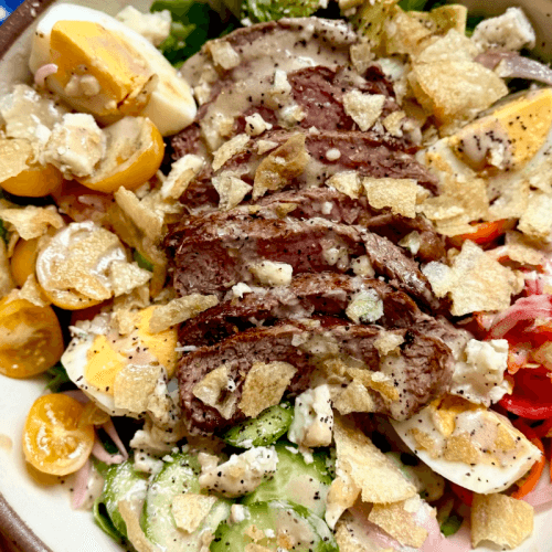 A delicious Steak Salad ready in just 15 Minutes, featuring sliced steak, cherry tomatoes, hard-boiled eggs, cucumbers, pickled onions, blue cheese, crushed potato chips, and a drizzle of Creamy Balsamic Dressing.