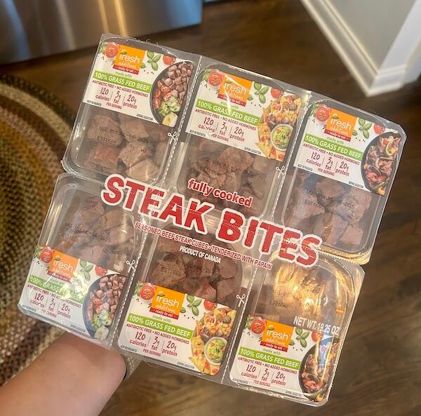 A hand holds a six-pack of Fresh brand fully cooked 100% grass-fed beef steak bites in clear plastic packaging—one of the popular Protein Picks at Costco.