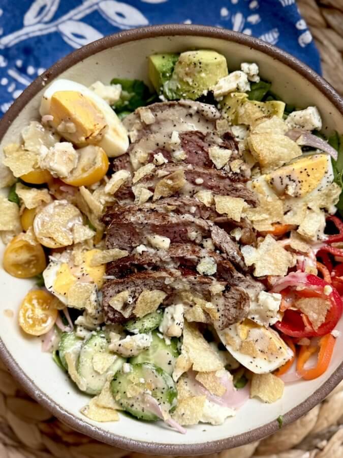 A vibrant Steak Salad featuring sliced steak, yellow cherry tomatoes, avocado, blue cheese, potato chips, cucumbers, and red peppers—all ready in just 15 Minutes—served with a blue patterned cloth in the background.