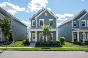 A two-story gray house with white trim at 6043 Gracious Dr, Franklin TN, featuring a front porch, central sidewalk, green lawn, and small trees—all showcased on a sunny day with blue sky and scattered clouds in this prime real estate listing.