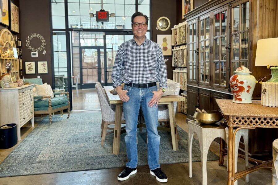 A man wearing glasses, a checkered shirt, and jeans stands smiling in the middle of Nashville Home Store, surrounded by stylish furniture and colorful home accessories.