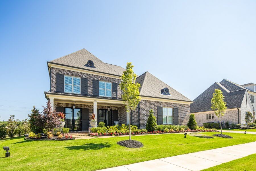Two-story brick house by Magnolia Homes with a covered front porch, manicured lawn, shrubs, and small trees on a sunny day. Adjacent houses are visible in the background—perfect for hosting your next Live Life Well Event.