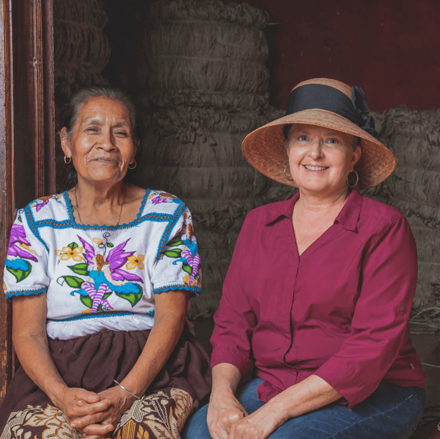 Two women sit side by side; one wears a colorful embroidered blouse, the other a maroon shirt and a wide-brimmed Tula Hat. Both are smiling, with bundles stacked in the background.