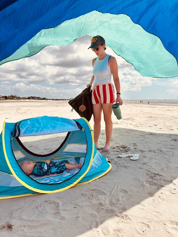 A woman stands on a sandy beach under a canopy, holding bags and a drink, while her baby relaxes in a small blue BabyQuip tent on the ground.