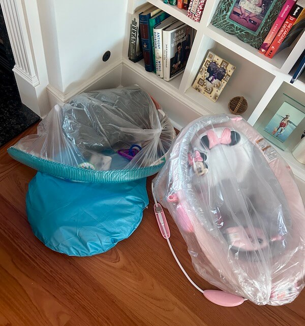 Two baby seats, one blue and one pink, are covered in clear plastic and placed on a wooden floor in front of a bookshelf with books and framed photos—perfect for your little ones' comfort, just like those offered by BabyQuip.
