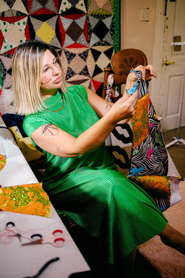 Kate Fusilier, a member of the Cosmin Collective, sits in a green dress and cuts fabric with scissors in a room adorned with a colorful quilt.