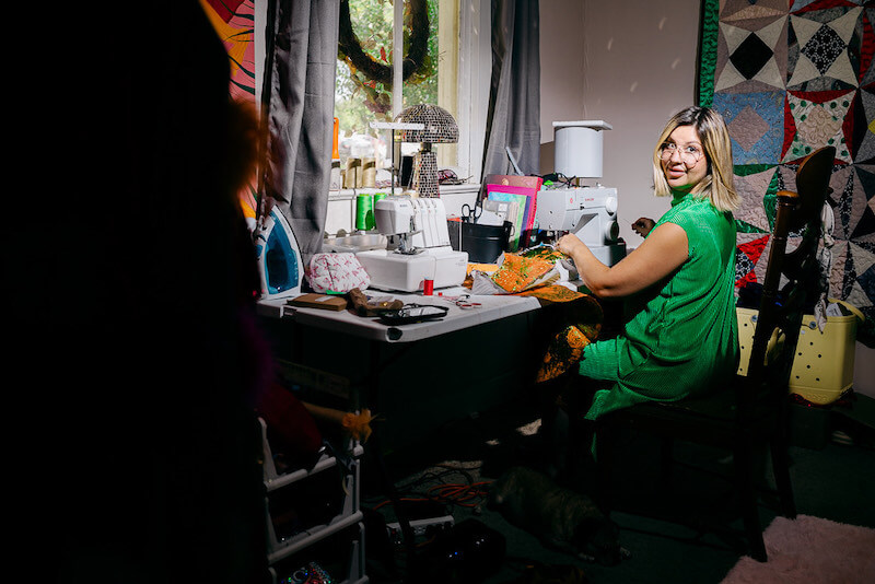 A woman in a green dress sits at a sewing machine in a well-lit room, surrounded by fabrics and sewing supplies. Kate Fusilier of Cosmin Collective smiles at the camera, capturing her creative spirit.