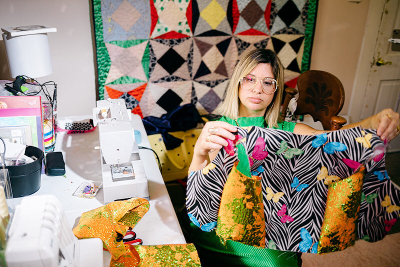 Kate Fusilier sits at a table with sewing machines, holding up a colorful fabric piece in front of a quilt hanging on the wall—a vibrant moment captured by Cosmin Collective.