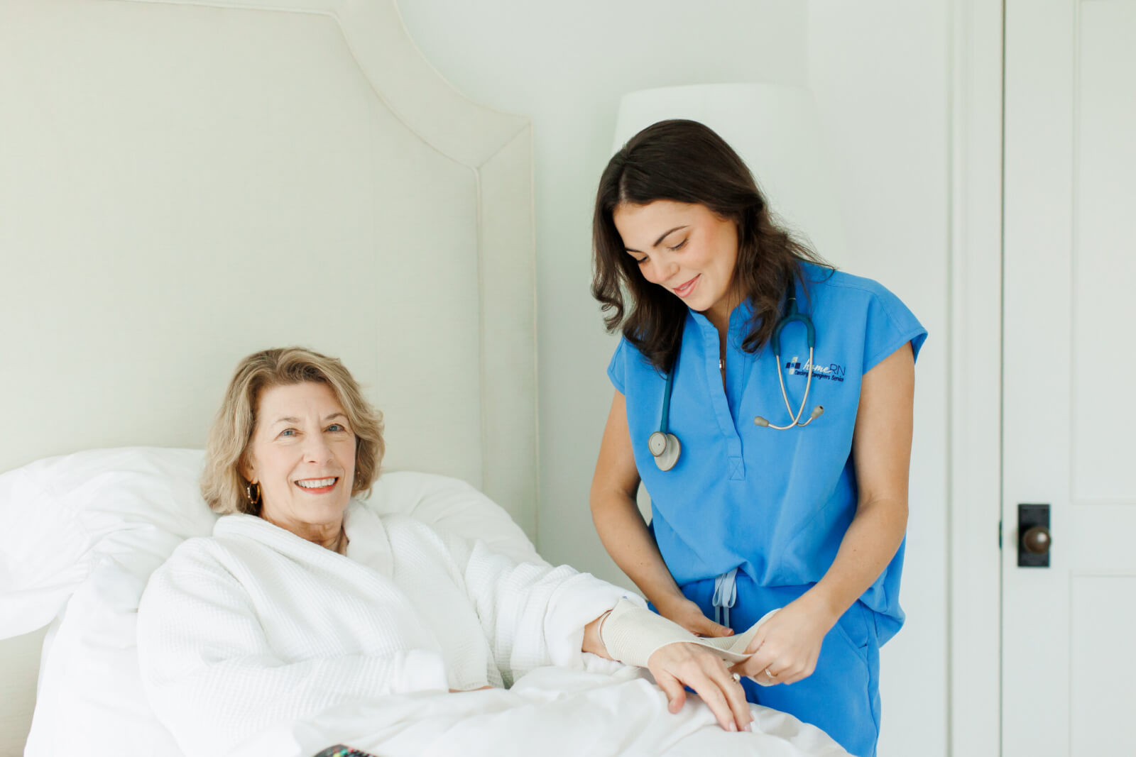 A nurse in blue scrubs from HomeRN Nashville checks the pulse of an older woman sitting up in bed, both smiling in a well-lit room—a warm example of compassionate home healthcare.