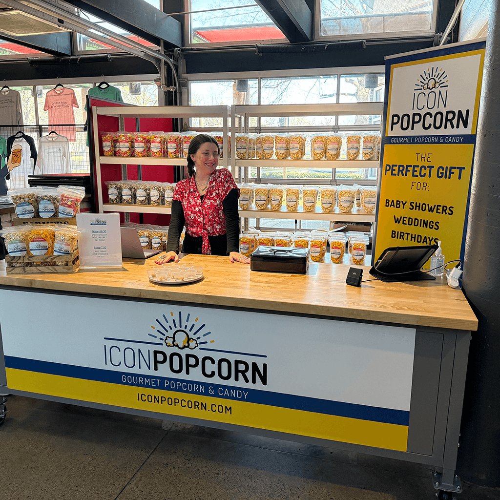 A woman stands behind a market booth displaying bags of Icon Popcorn, with a sign reading "ICON POPCORN" and a banner promoting their gourmet popcorn as the perfect gift for events.