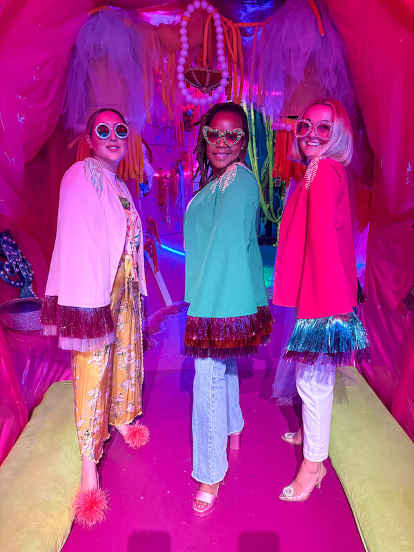 Three women from the Cosmic Collective stand side by side in a colorful, neon-lit room, wearing large sunglasses, bright jackets with fringe, and stylish outfits.