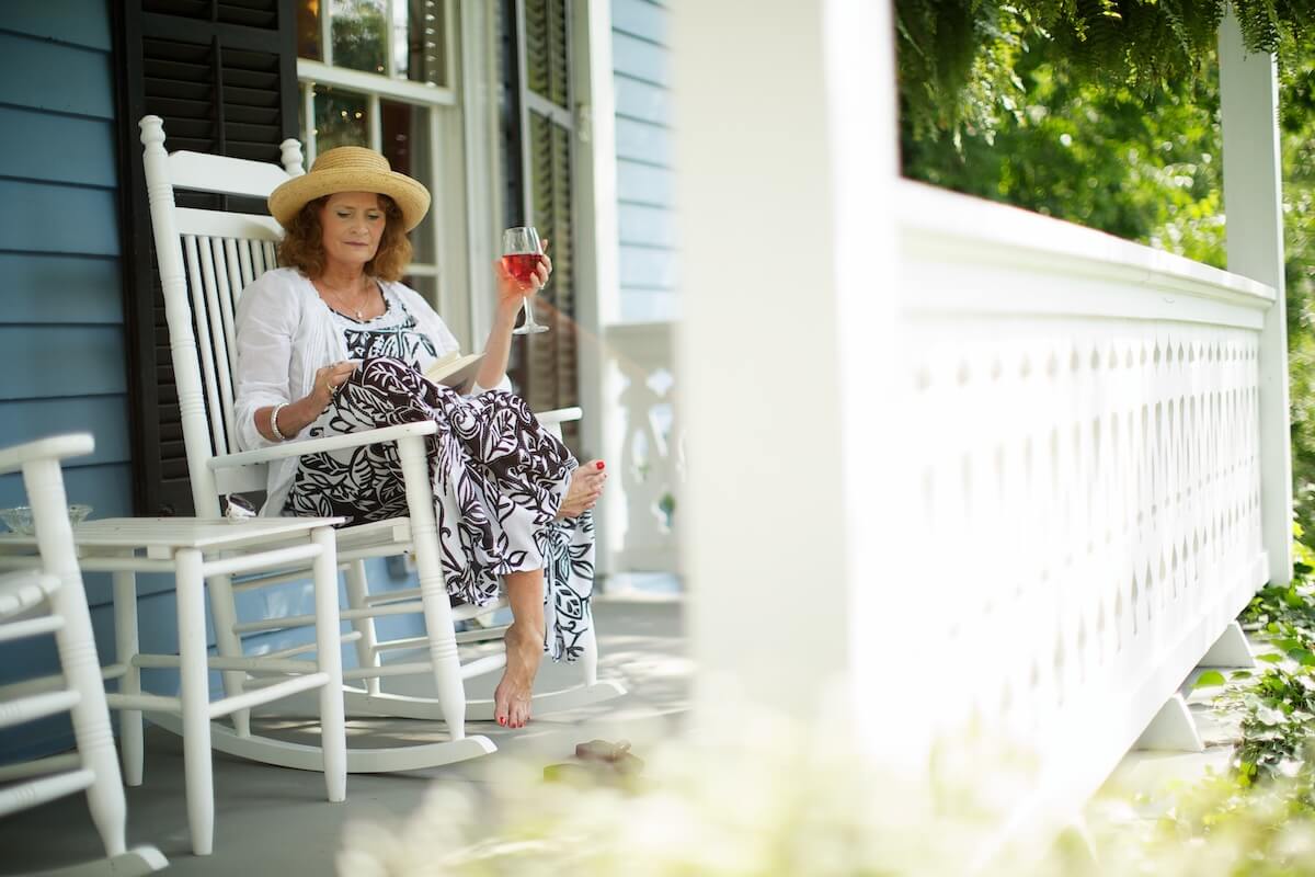 A woman in a hat sits on a white rocking chair on a porch, reading a book and holding a glass of pink wine—embracing the charm found in the best towns in Virginia.