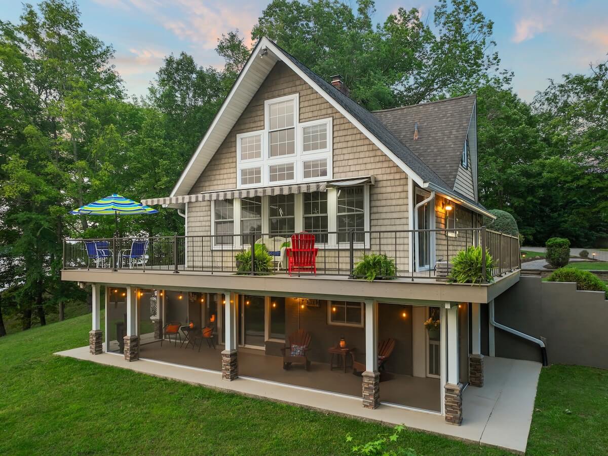 Two-story house with large windows, upper deck with outdoor seating, and covered lower patio, surrounded by grass and trees—perfect for relaxing weekend getaways in Virginia.