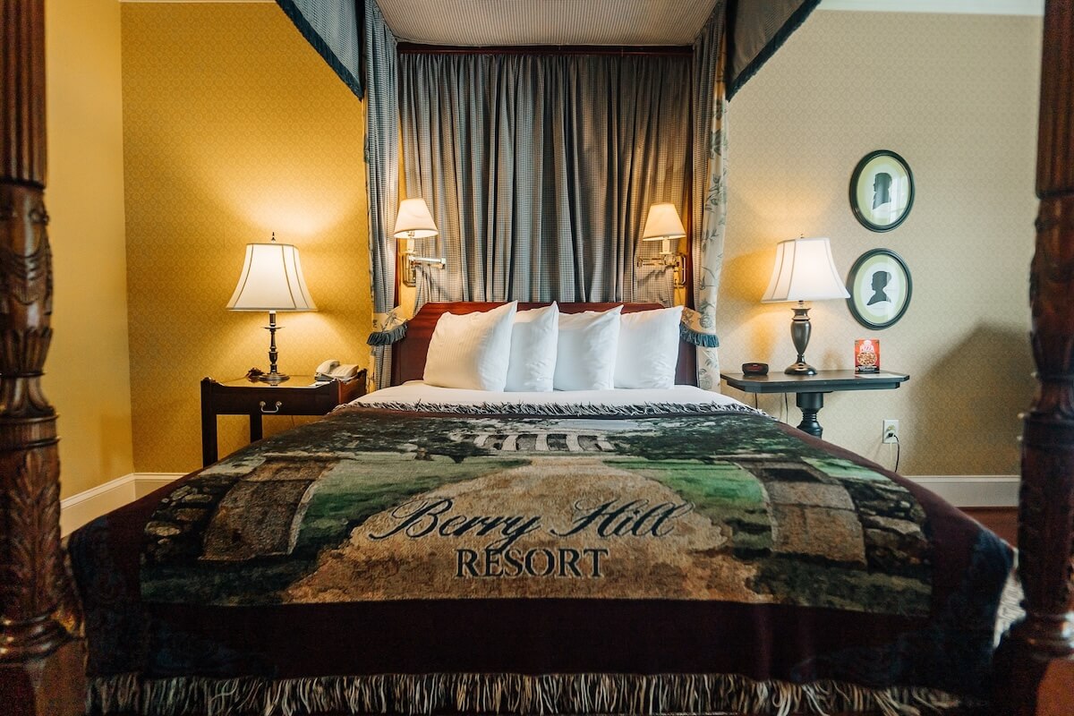Four-poster bed with canopy, five pillows, and lamps on bedside tables; a "Berry Hill Resort" blanket covers the bed. Two silhouette portraits hang on the wall—perfect for relaxing weekend getaways in Virginia.