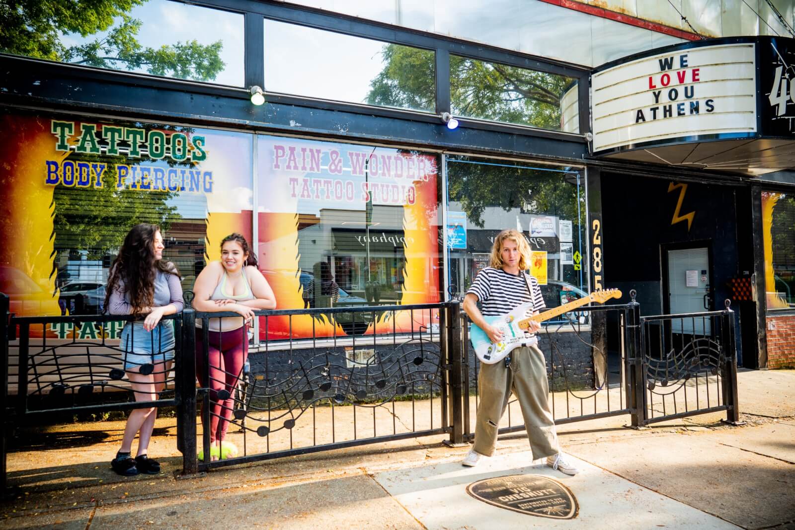 Three people stand outside a tattoo and piercing shop in one of the lively SEC Towns, with one playing an electric guitar. A marquee sign above reads "WE LOVE YOU ATHENS.