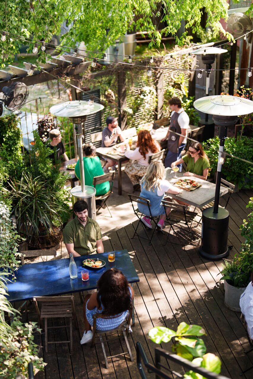 People are dining and socializing at outdoor tables on a wooden patio, surrounded by greenery and garden heaters—a lively scene reminiscent of gatherings in SEC Towns during the daytime.