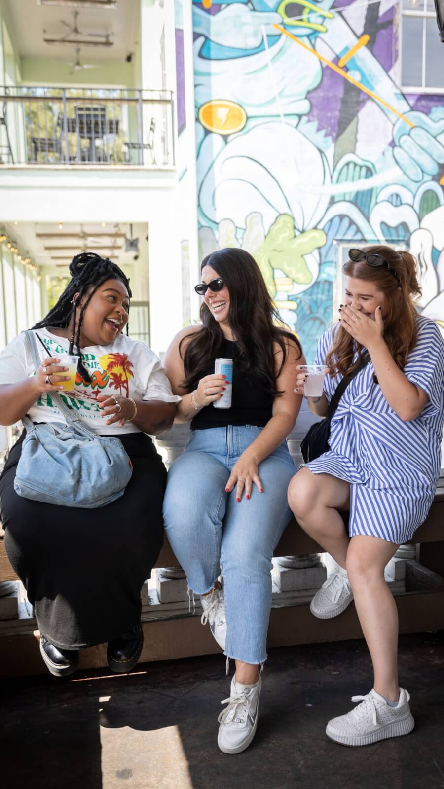Three women sit on a bench outdoors in one of the vibrant SEC Towns, laughing and holding drinks in front of a colorful mural.