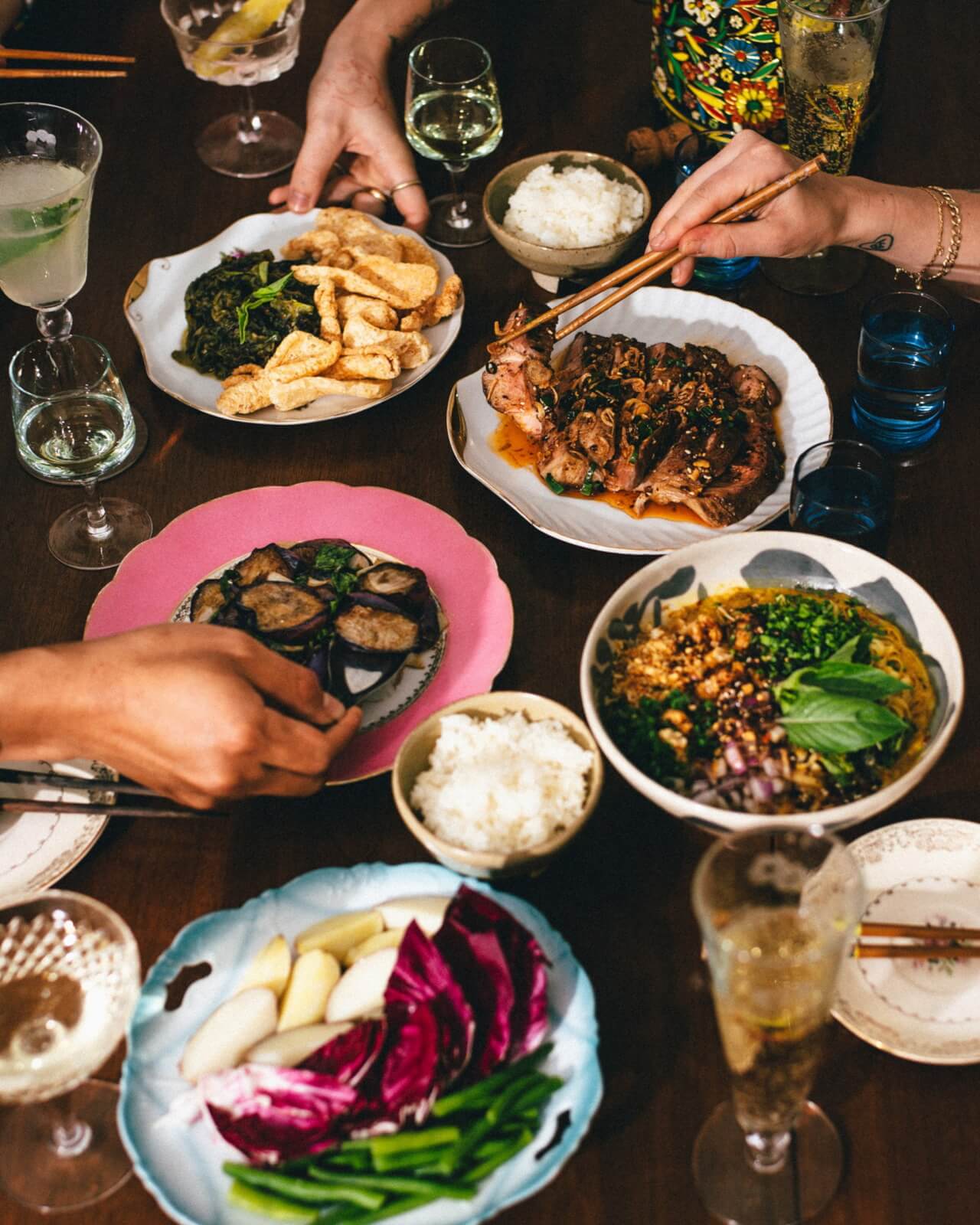 A table set with various Asian dishes, bowls of rice, glasses of drinks, and three people using chopsticks to serve themselves.